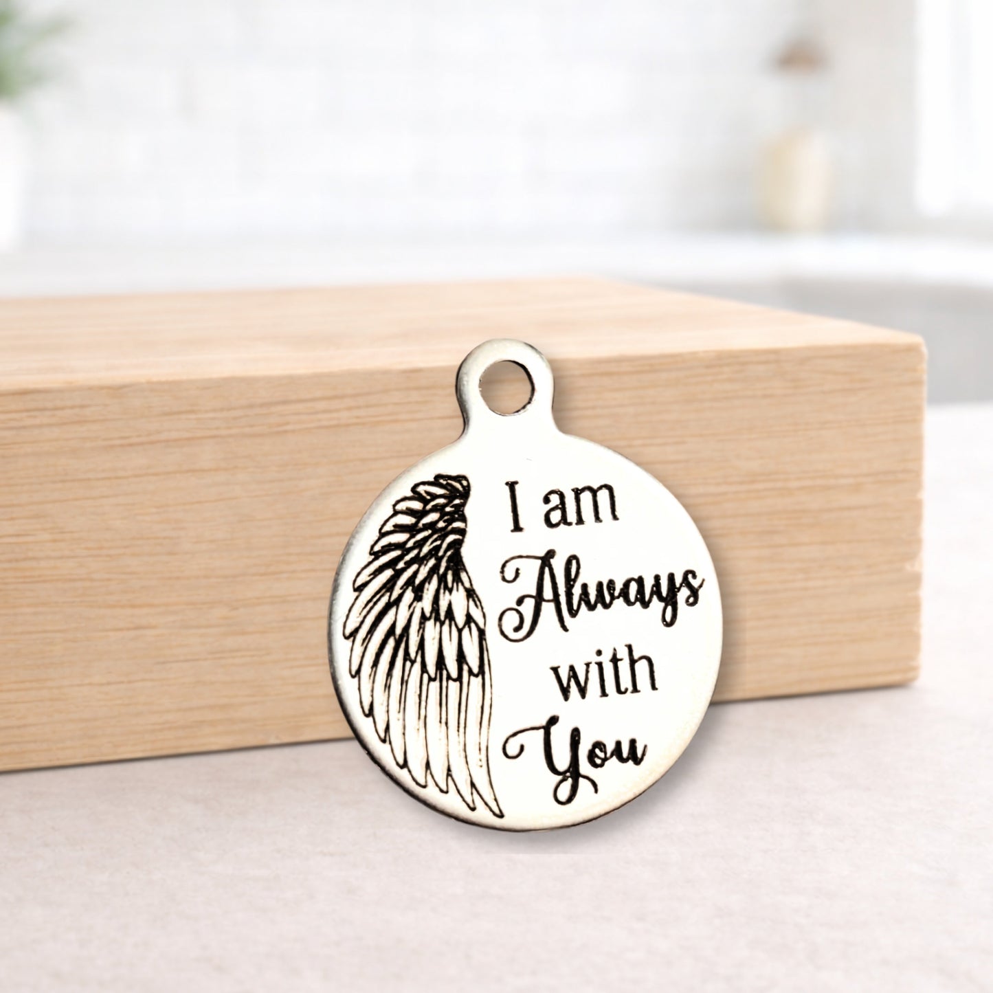 Metal pendant with angel wing design and 'I am Always with You' text on a wooden block.