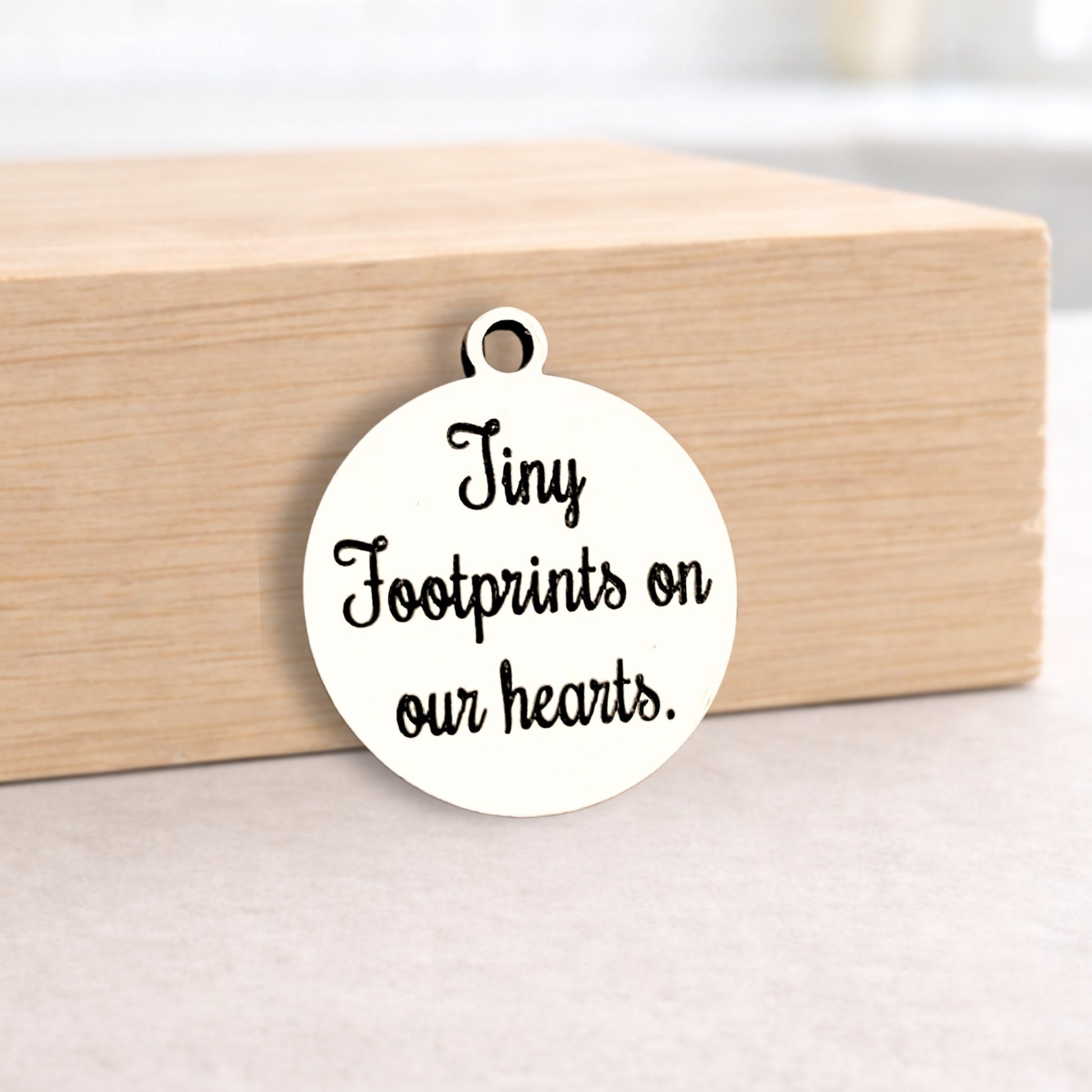 Round metal pendant with 'Tiny Footprints on our hearts.' text on a wooden block.