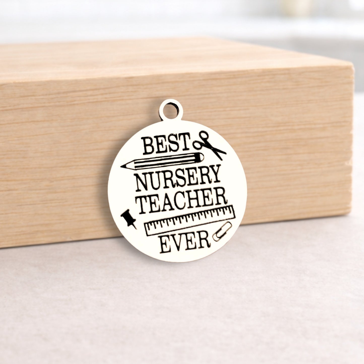 Round metal pendant with 'Best Nursery Teacher Ever' text on a wooden block.