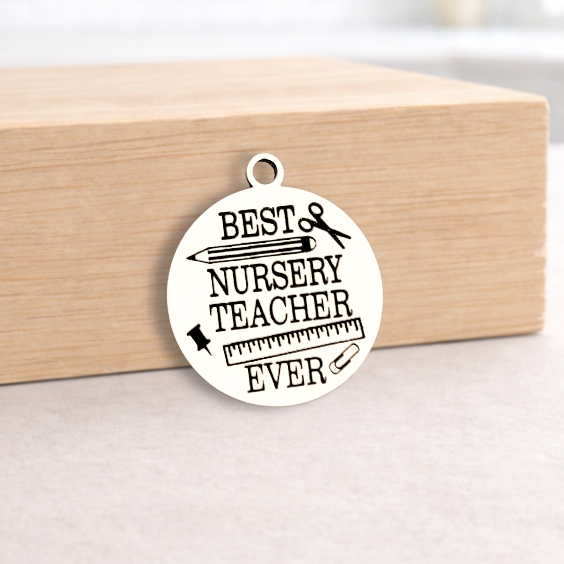 Round metal pendant with 'Best Nursery Teacher Ever' text on a wooden block.