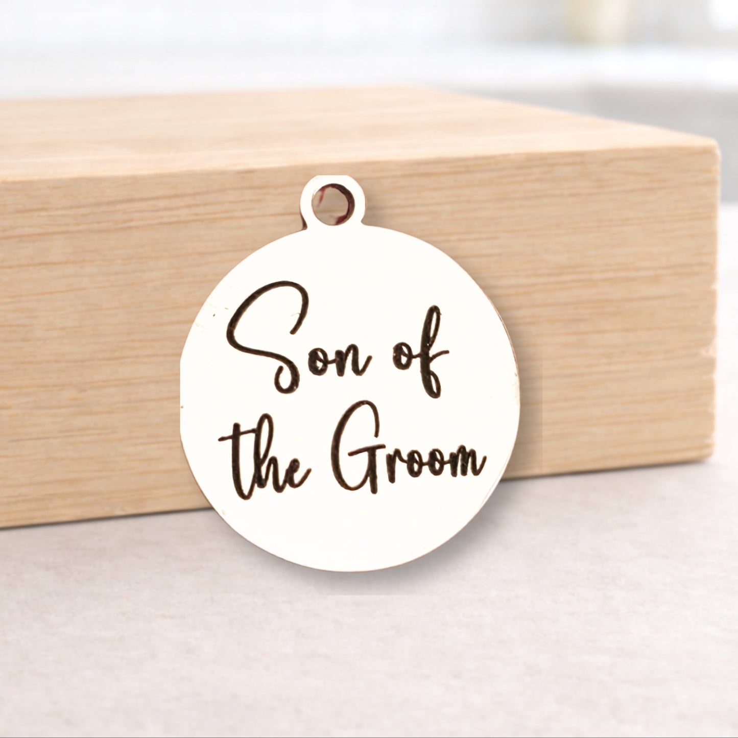 Round white pendant with 'Son of the Groom' text on a wooden block.