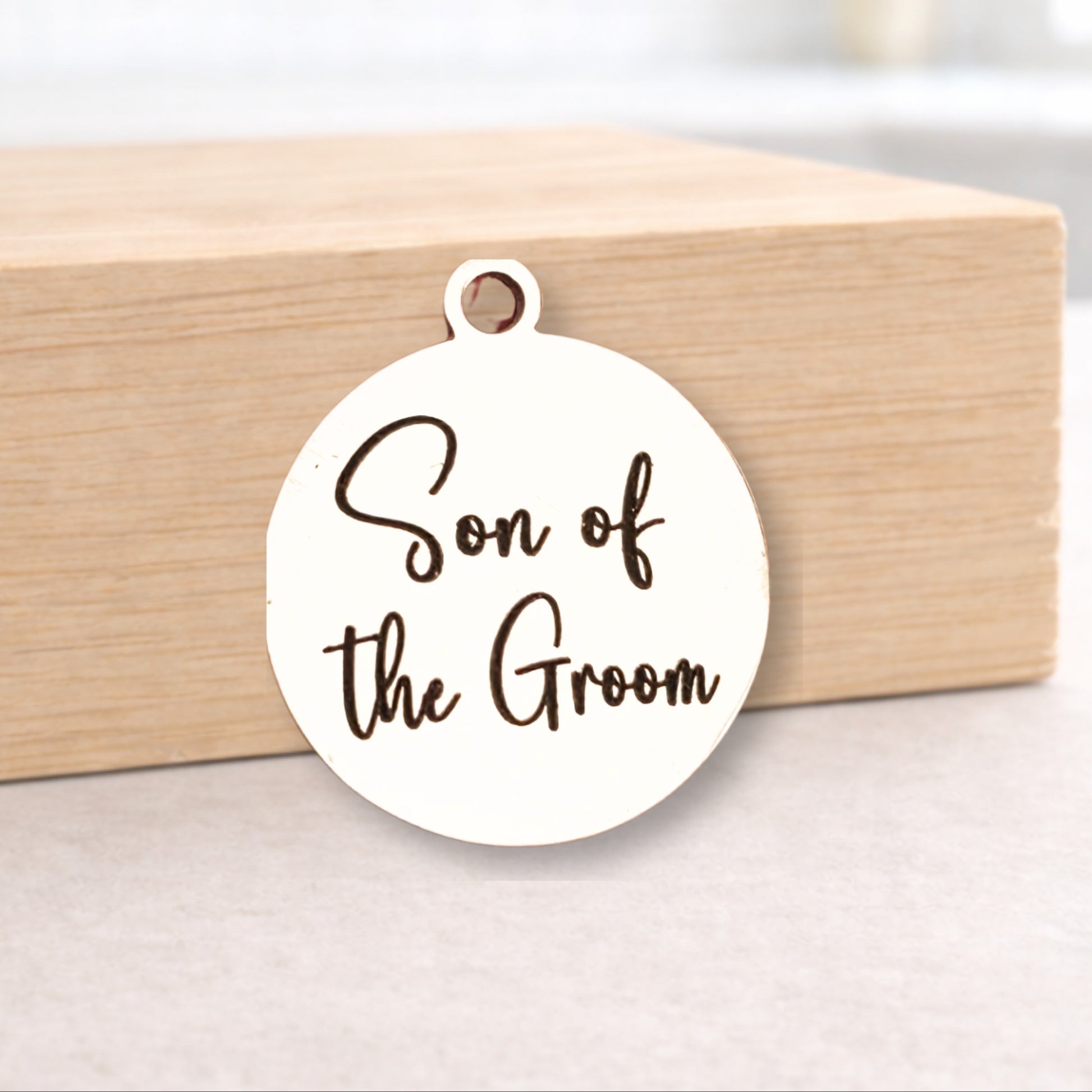 Round white pendant with 'Son of the Groom' text on a wooden block.