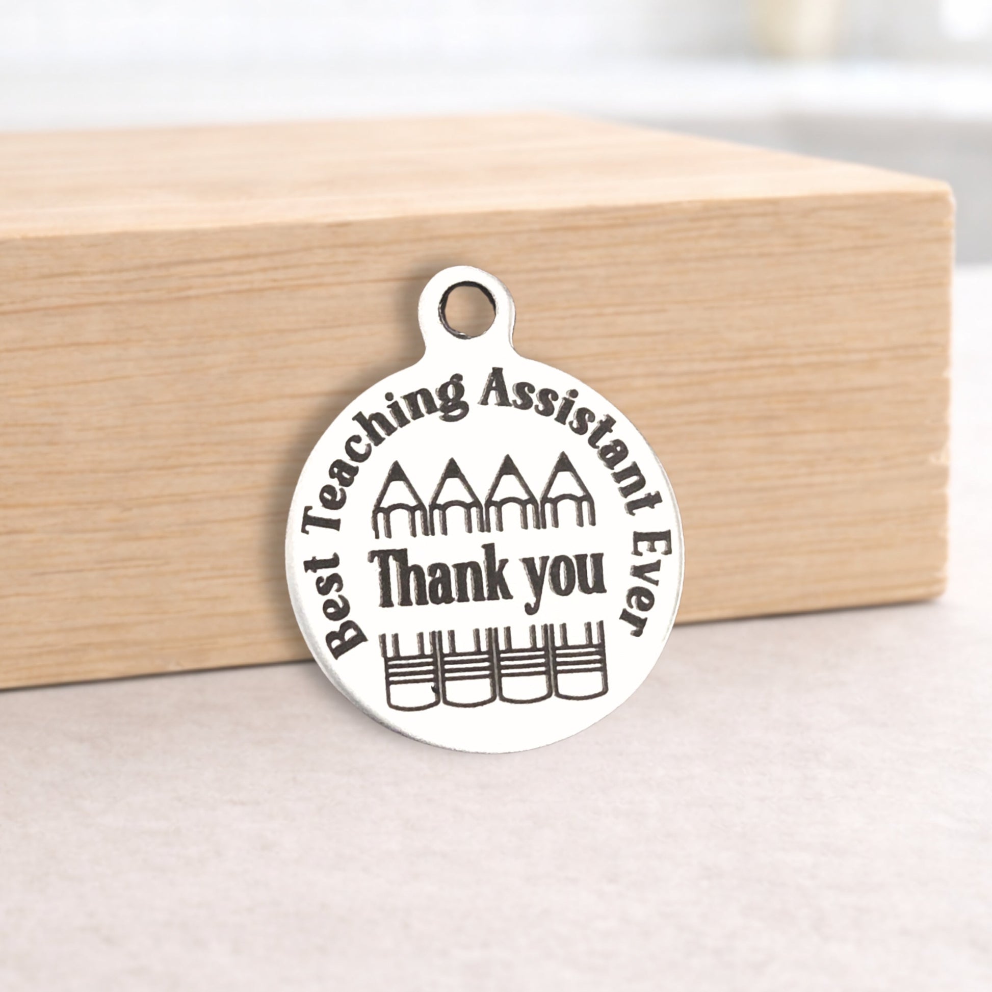Silver pendant with 'Best Teaching Assistant Ever Thank you' text on a wooden block.