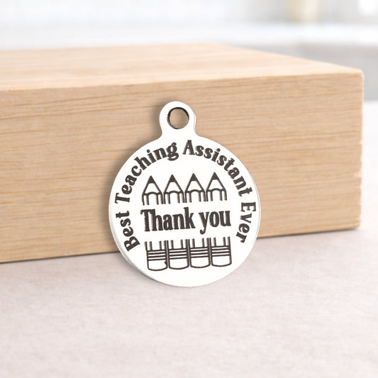 Silver pendant with 'Best Teaching Assistant Ever Thank you' text on a wooden block.