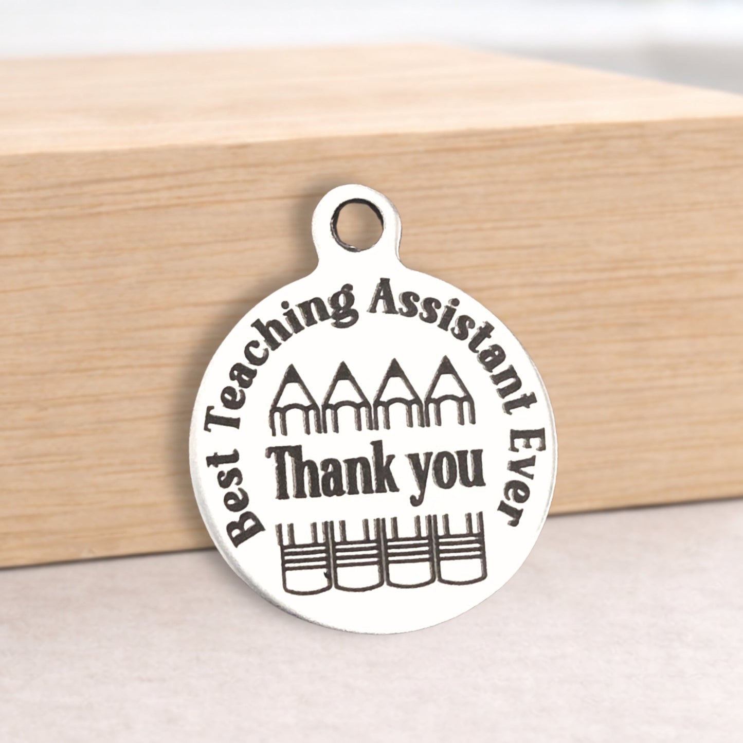 Silver pendant with 'Best Teaching Assistant Ever Thank you' text on a wooden block.
