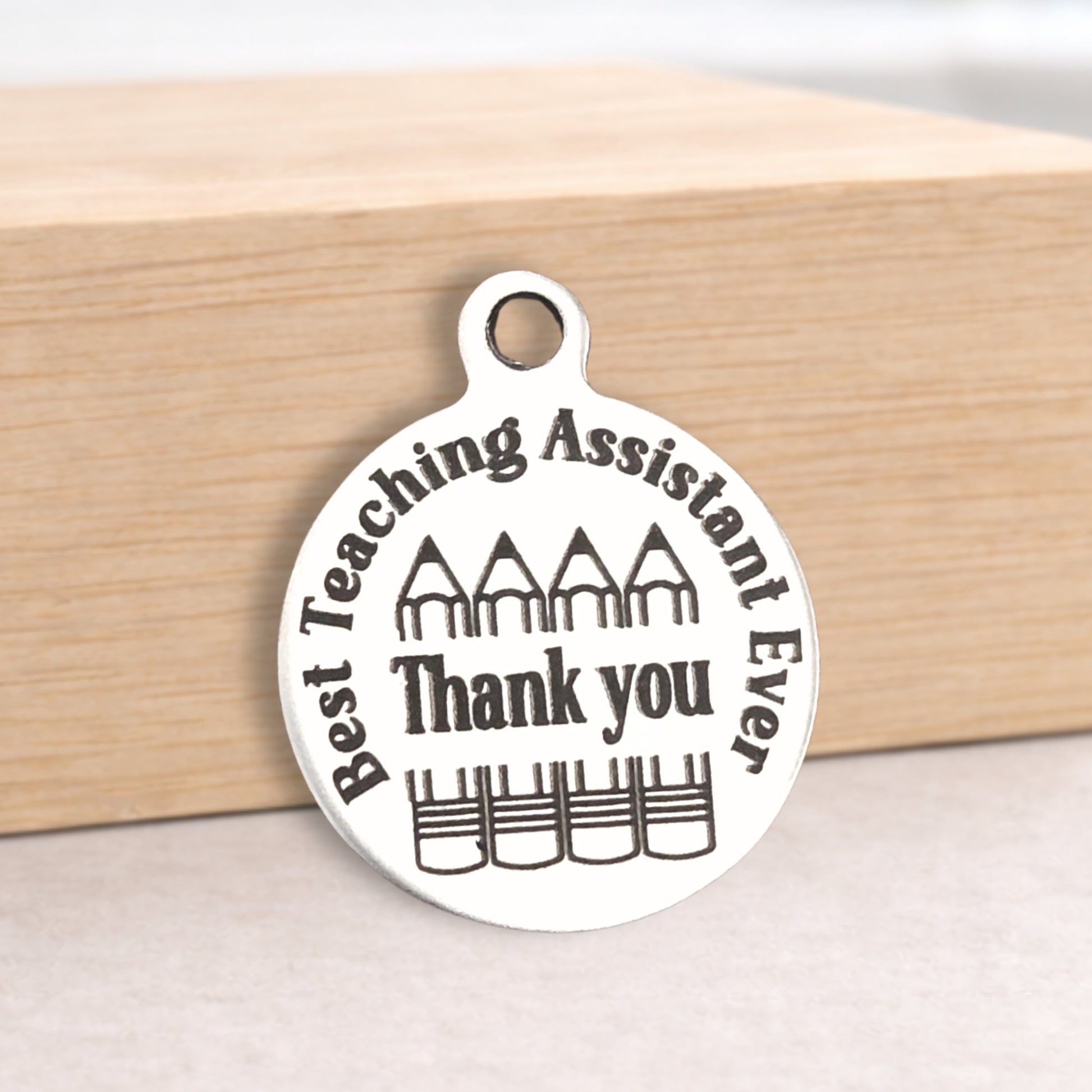 Silver pendant with 'Best Teaching Assistant Ever Thank you' text on a wooden block.