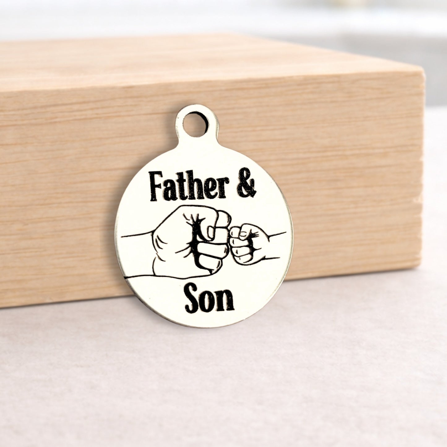Circular metal pendant with 'Father & Son' text and fist bump design on a wooden block.