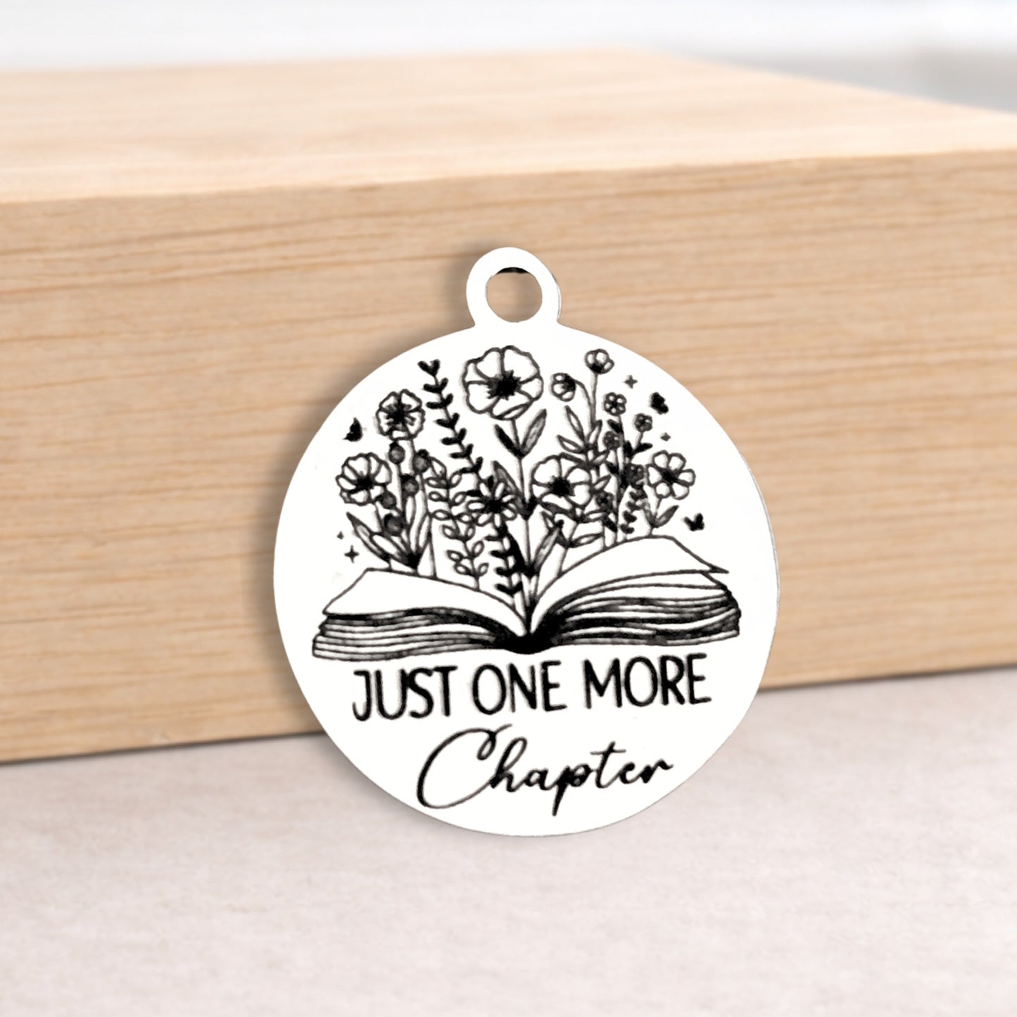 Circular pendant with 'Just One More Chapter' text and floral design on a wooden block.