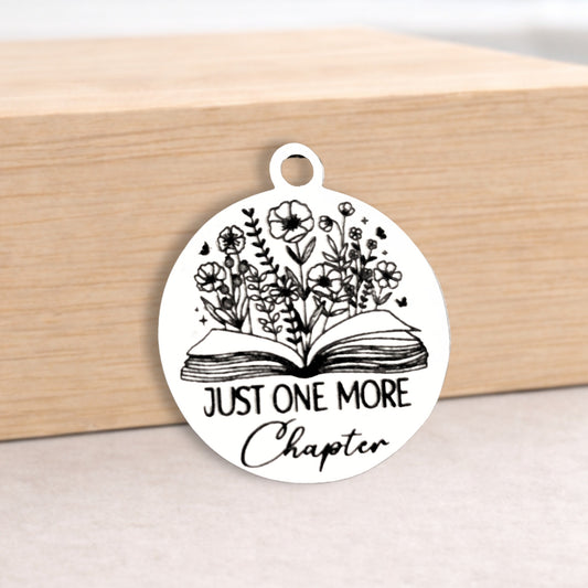 Circular pendant with 'Just One More Chapter' text and floral design on a wooden block.