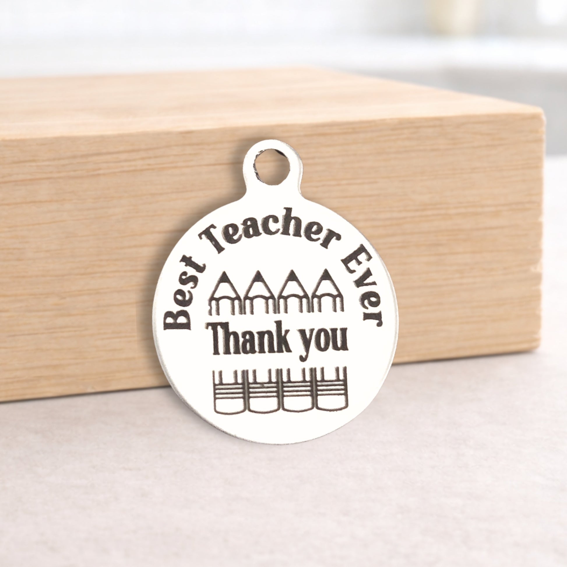 Round metal pendant with 'Best Teacher Ever Thank you' text on a wooden block.