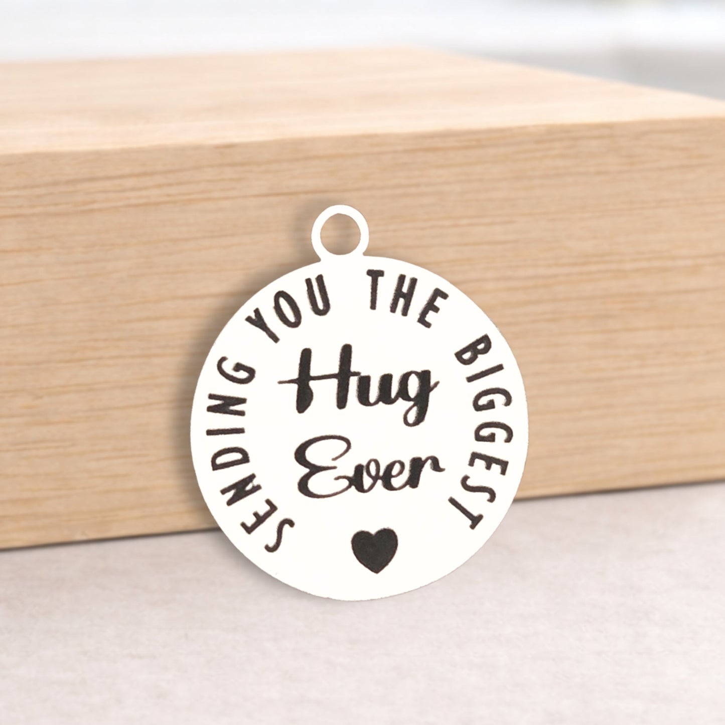 Round metal pendant with 'Sending you the biggest hug ever' text on a wooden block.