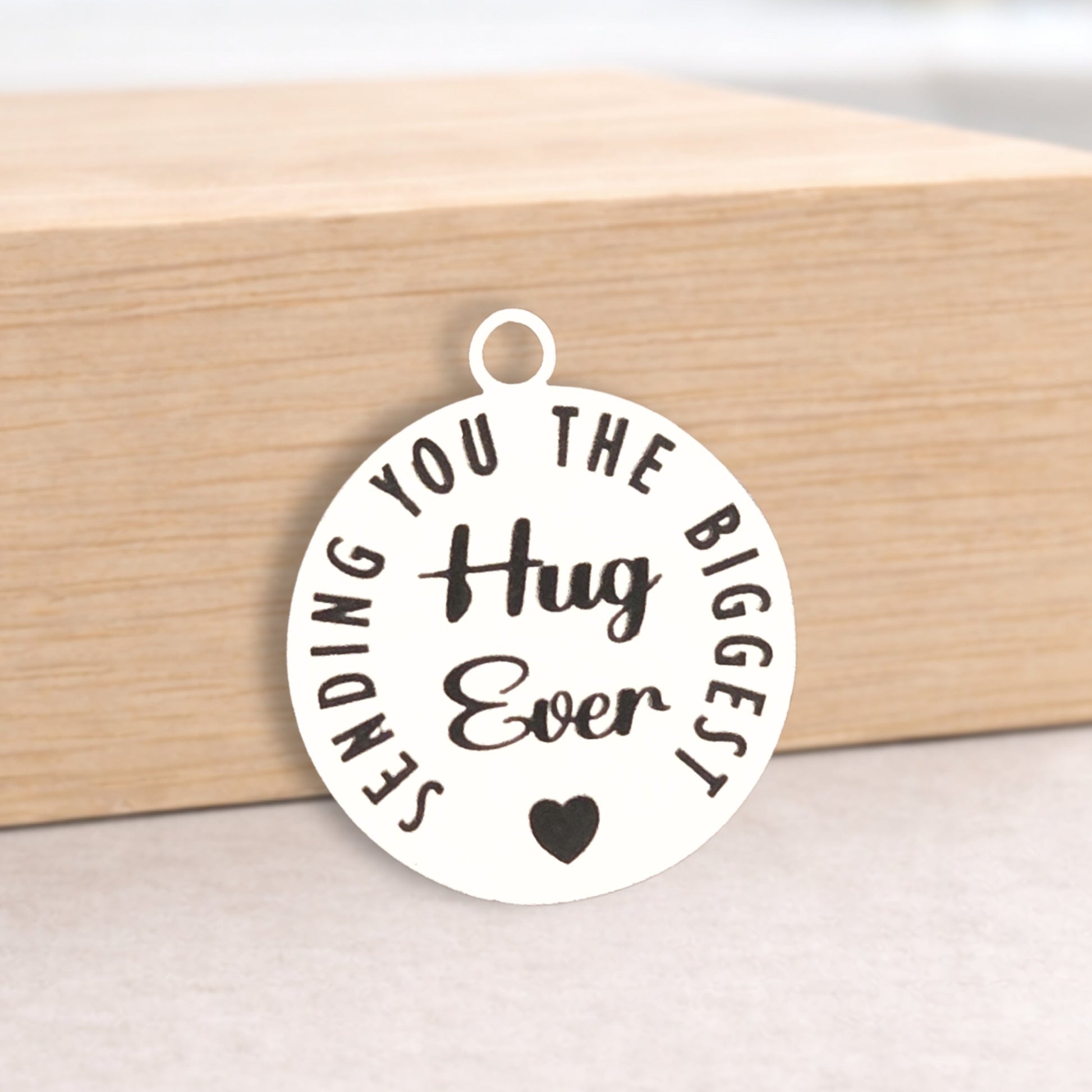 Round metal pendant with 'Sending you the biggest hug ever' text on a wooden block.