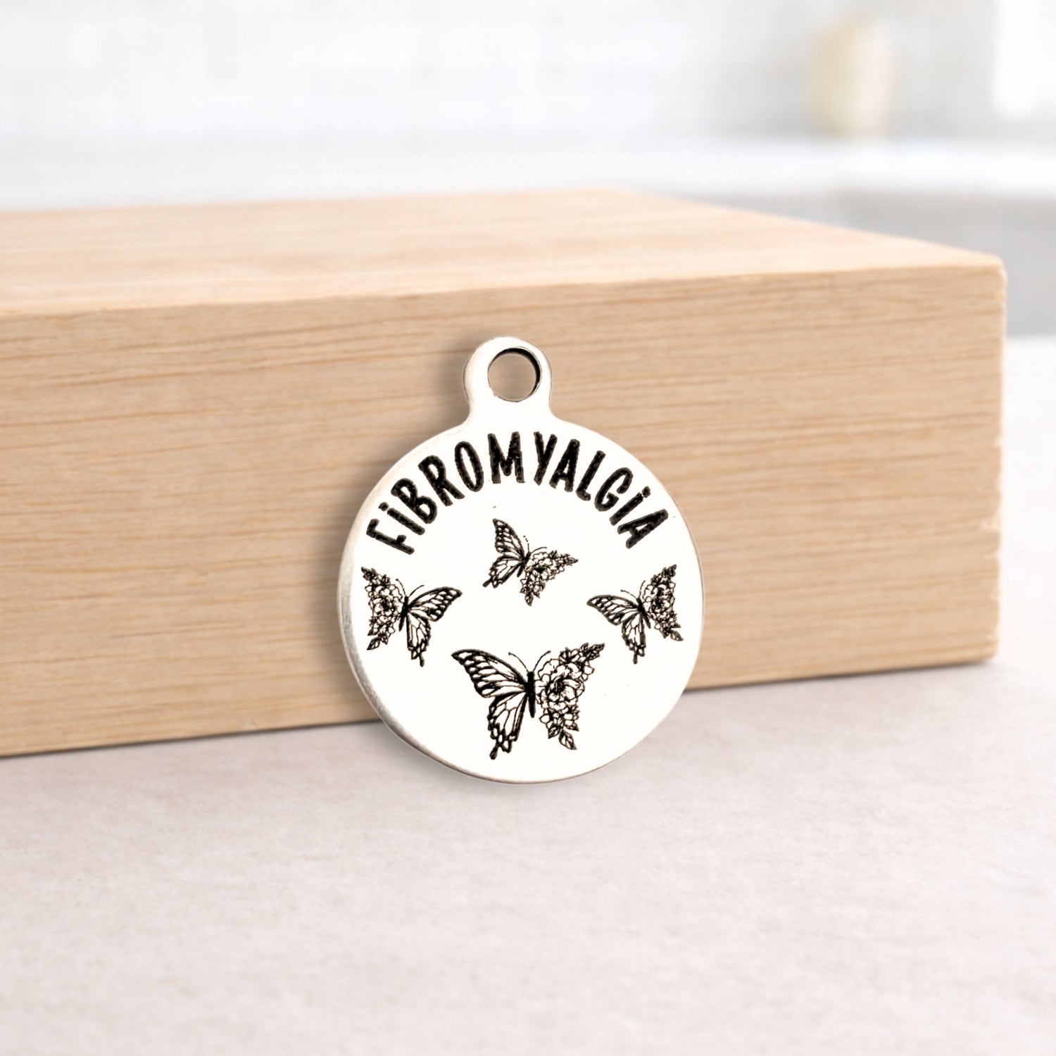 Silver pendant with 'FIBROMYALGIA' and butterfly designs on a wooden block.