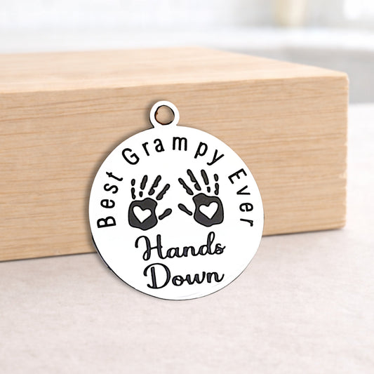 Round pendant with 'Best Grampy Ever Hands Down' text and handprints on a wooden surface.