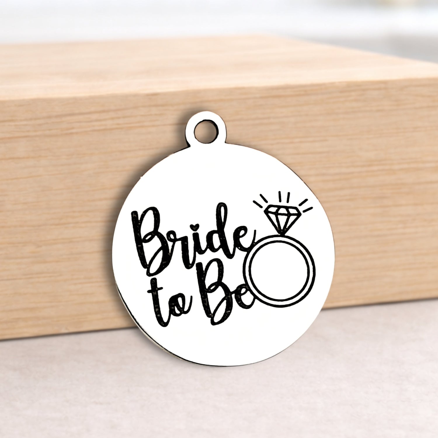 Round metal pendant with 'Bride to Be' text and ring illustration on a wooden block.