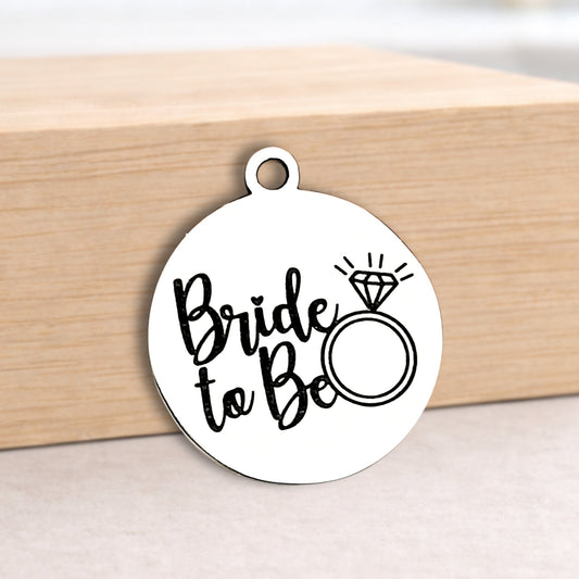 Round metal pendant with 'Bride to Be' text and ring illustration on a wooden block.