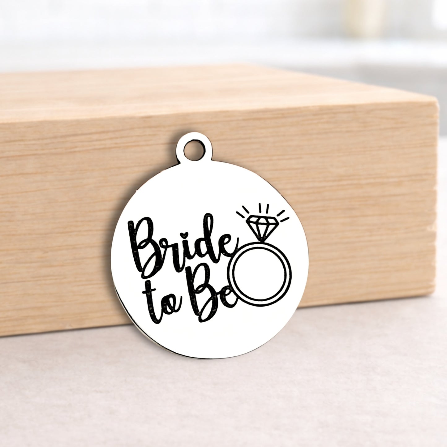 Round metal pendant with 'Bride to Be' text and ring illustration on a wooden block.