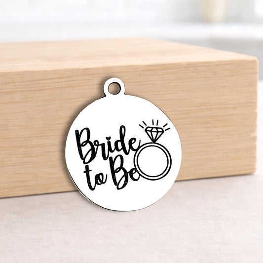 Round metal pendant with 'Bride to Be' text and ring illustration on a wooden block.