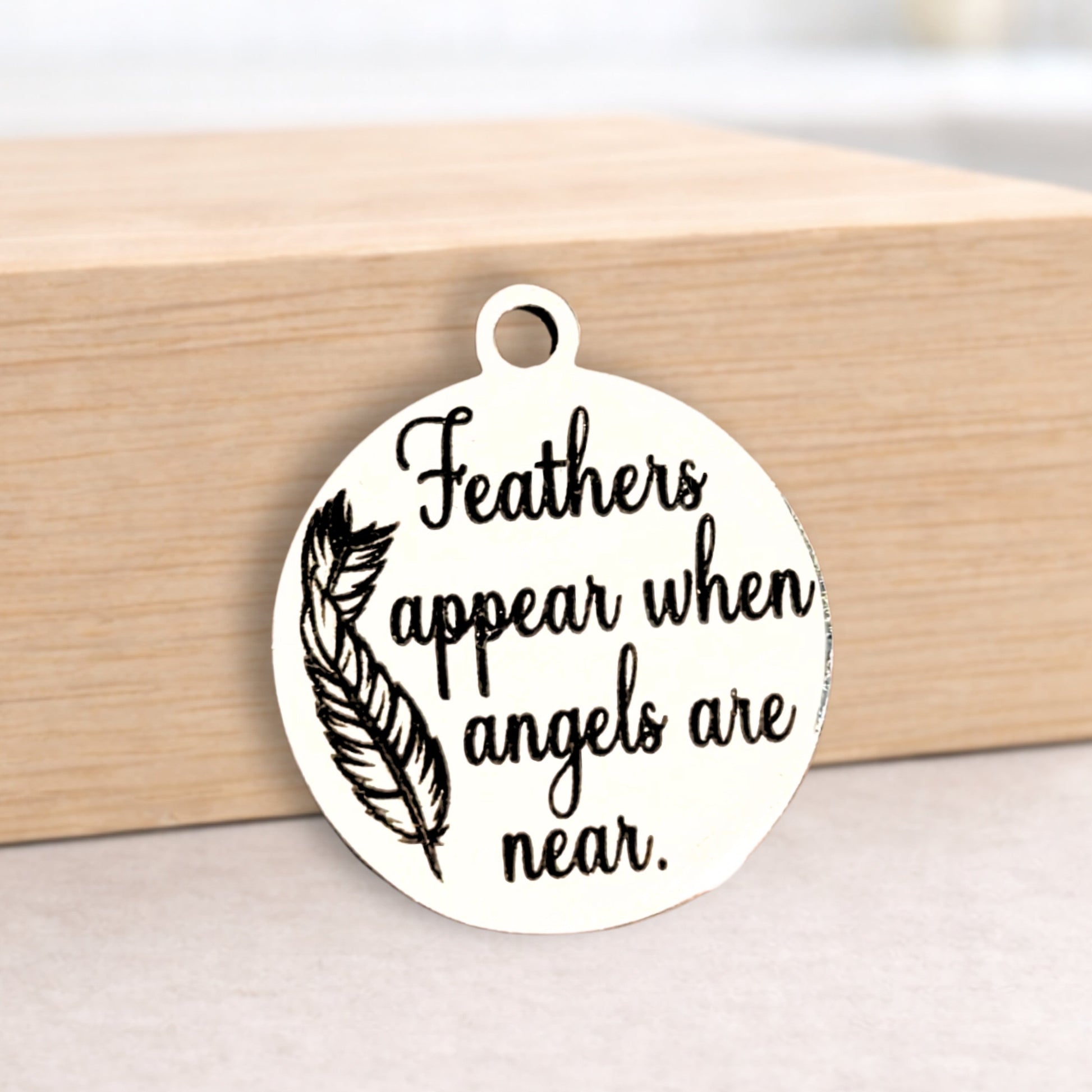 Round metal pendant with text 'Feathers appear when angels are near' on a wooden block.