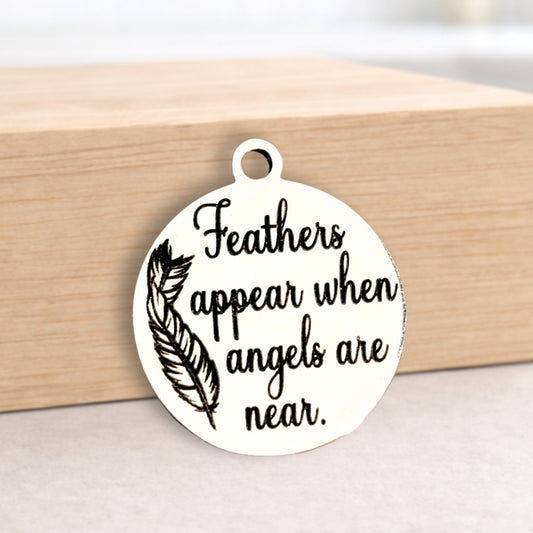 Round metal pendant with text 'Feathers appear when angels are near' on a wooden block.