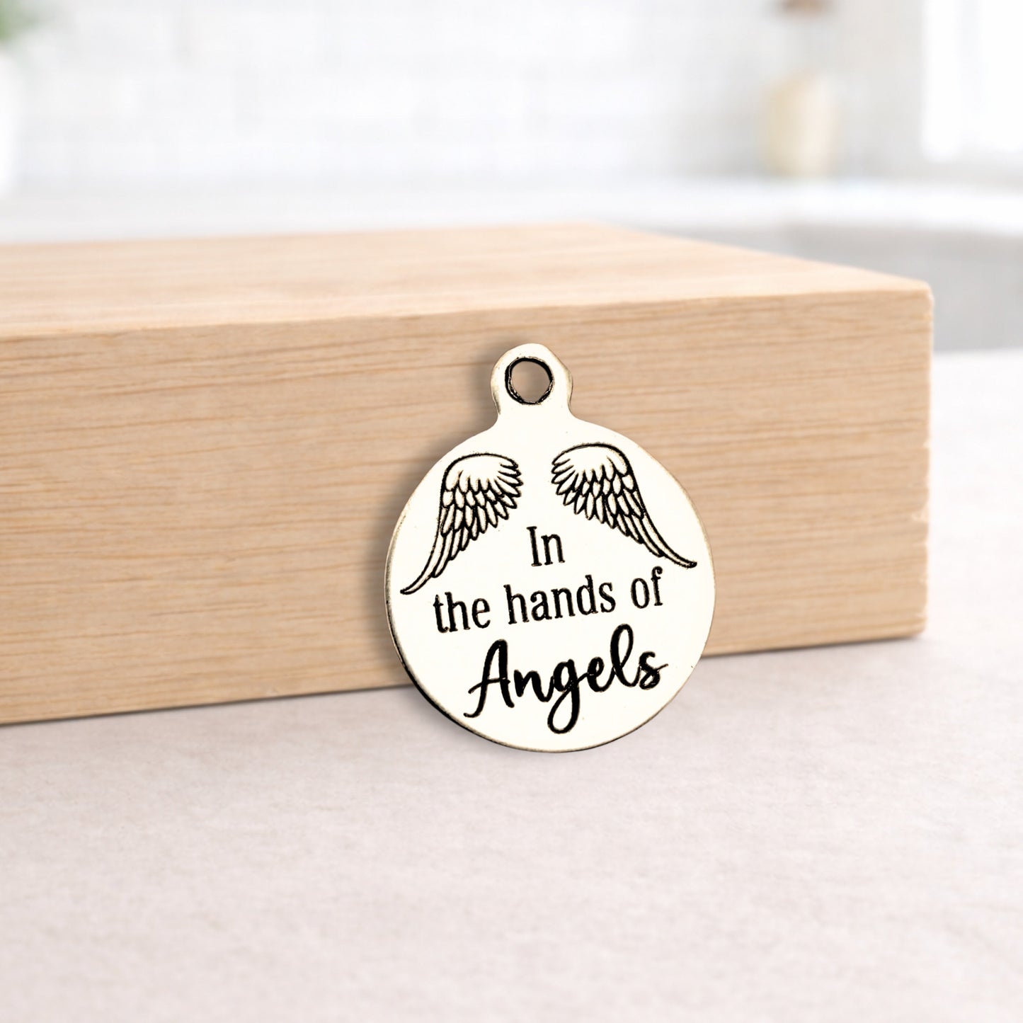 Metal pendant with 'In the hands of Angels' text and wings on a wooden block.