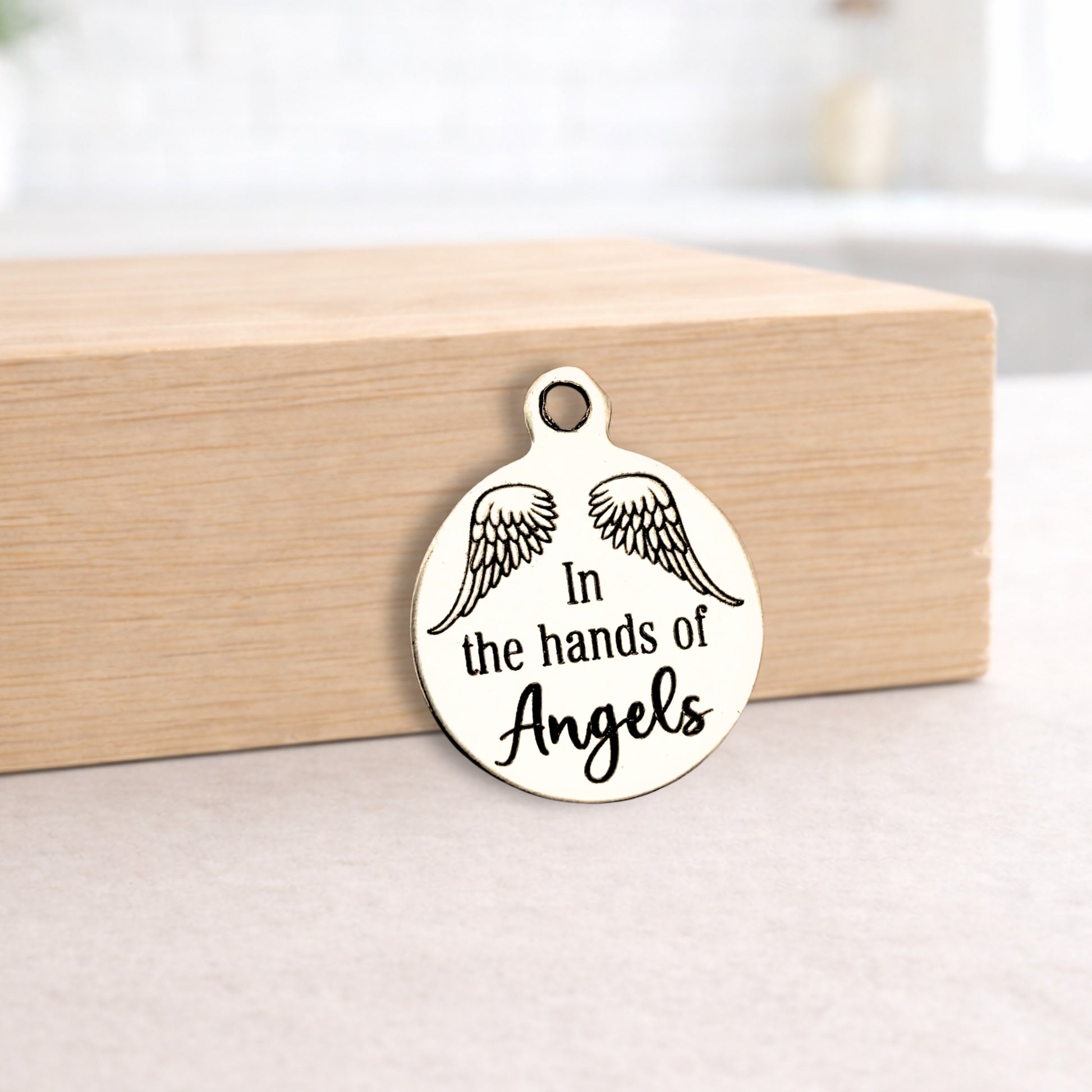 Metal pendant with 'In the hands of Angels' text and wings on a wooden block.