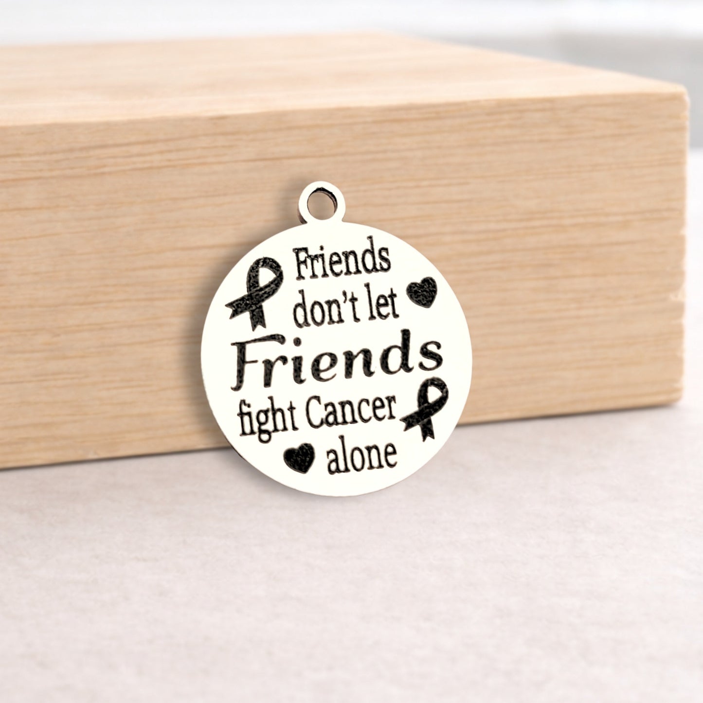 White pendant with black text and cancer awareness ribbons on a wooden block.