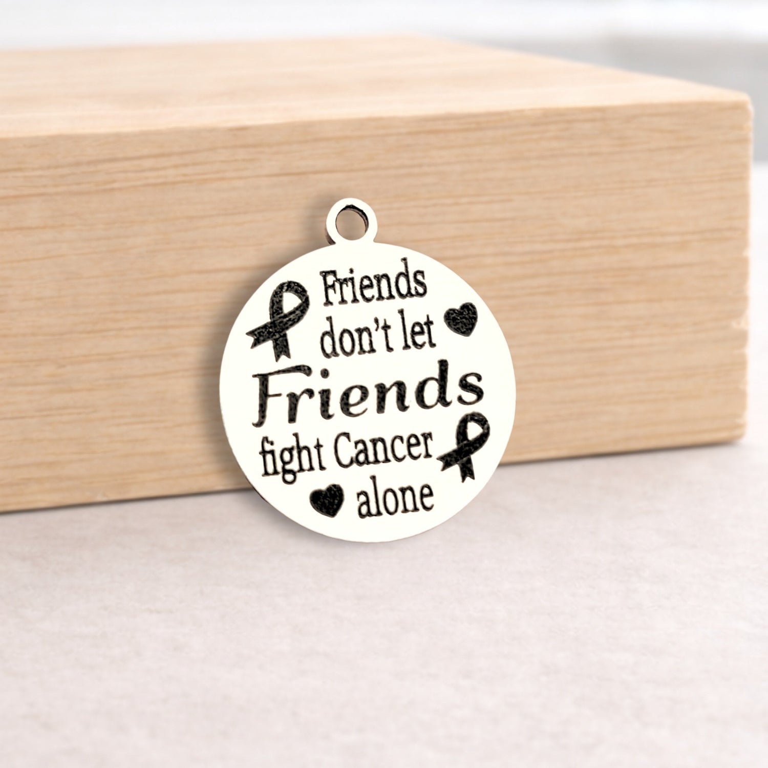 White pendant with black text and cancer awareness ribbons on a wooden block.