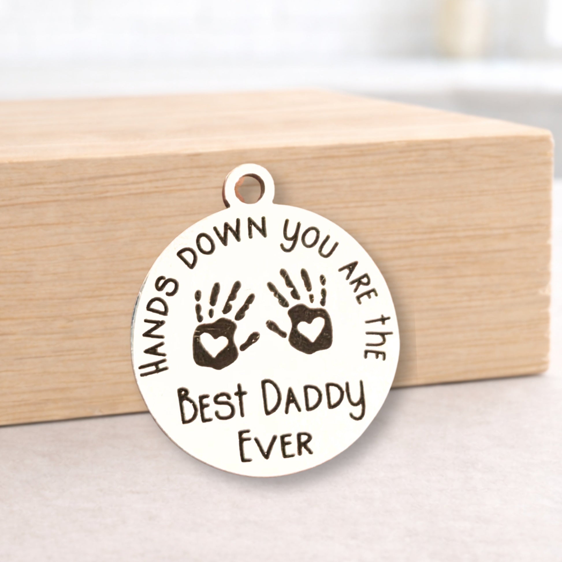 Round metal pendant with engraved text and handprints on a wooden block.