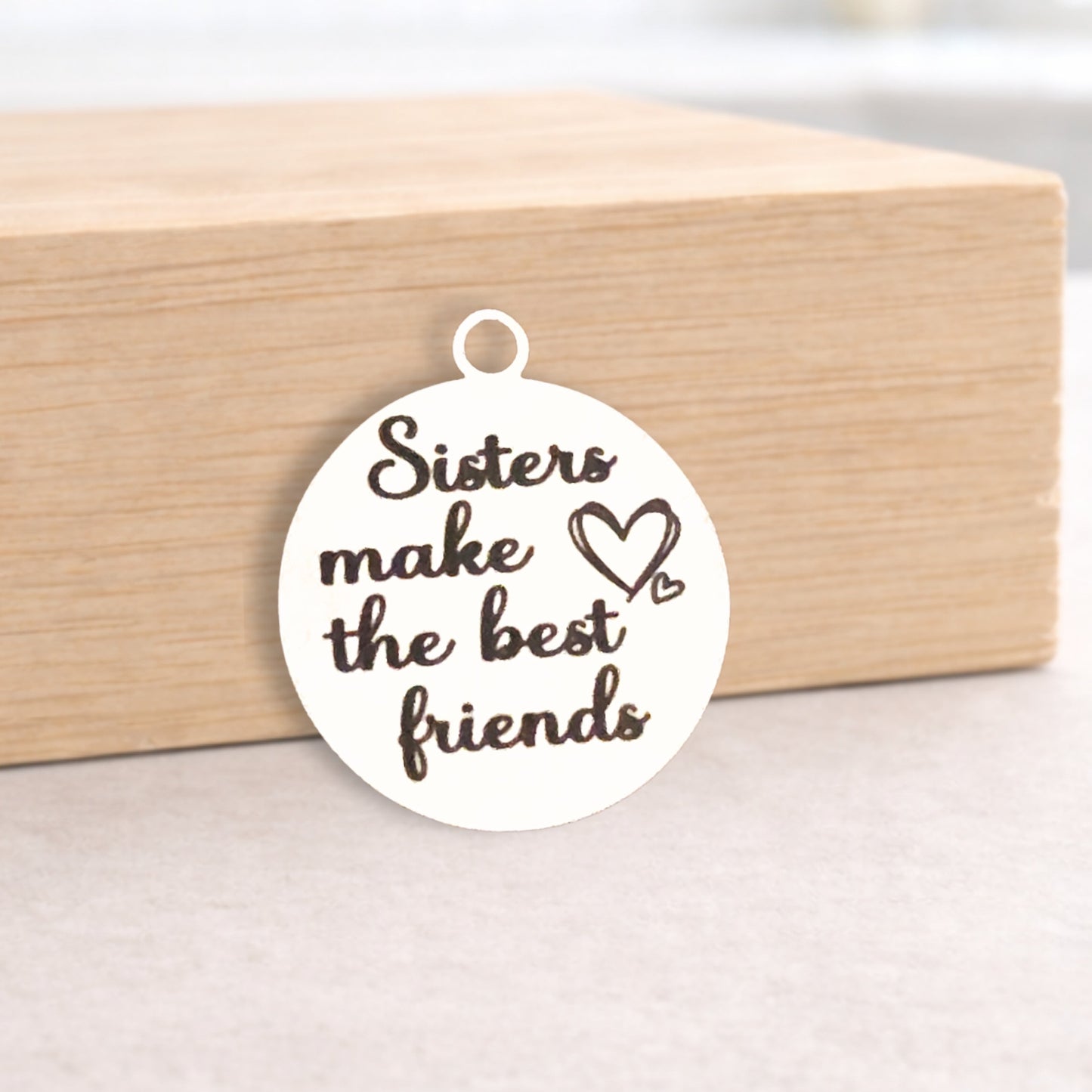 Round white pendant with 'Sisters make the best friends' text on a wooden block.
